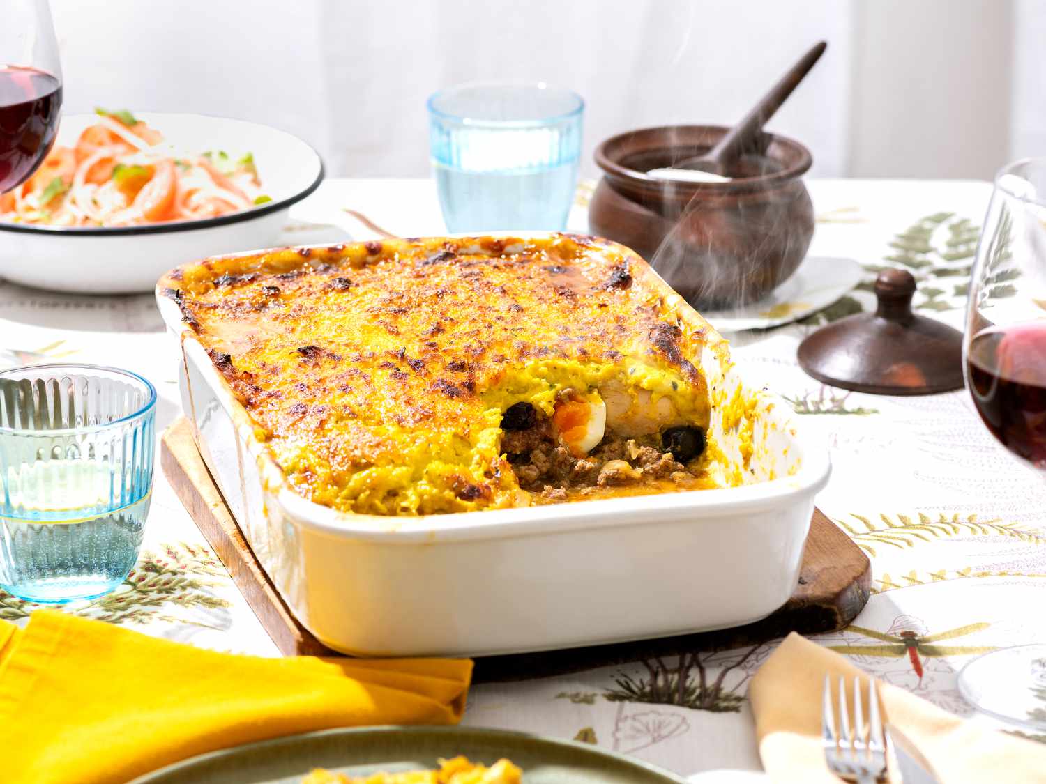 Horizontal side view of Pastel De Choclo with a serving taken out of it. On the table with the tray is glasses of wine, water, a small dish of salt