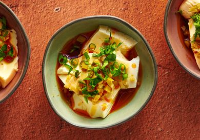 An irregularly round glazed ceramic bowl holding silken tofu with soy sauce and chili oil. There are two additional bowls, one on the right periphery of the image, one on the left periphery.