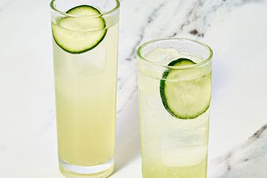 Two glasses with cucumber garnished beverages on a marble surface