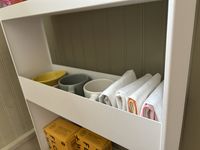 A closeup of a shelf of a white kitchen storage rack, with napkins and mugs on it