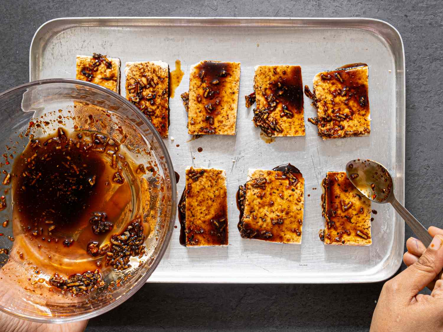 Marinated tofu on a baking tray with sauce being added by a spoon