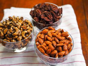Three glass dishes of spiced nuts on a kitchen towel on a table. The flavors are smoky candied almonds, Mexican spiced chocolate pecans, and olive-rosemary cashews.