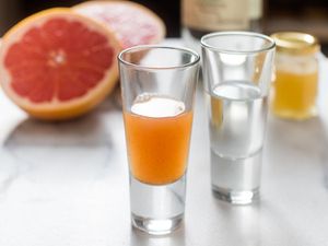 Closeup of a shot glass of grapefruit sangrita with chipotle and honey, flanked by another shot glass full of silver tequila. A halved grapefruit is in the blurred background.