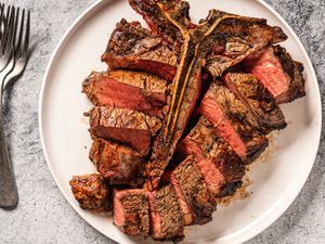 A t-bone (porterhouse) steak on a white plate, with the sliced meat placed around the bone. On the lefthand side of the image are several metal forks stacked on top of each other.