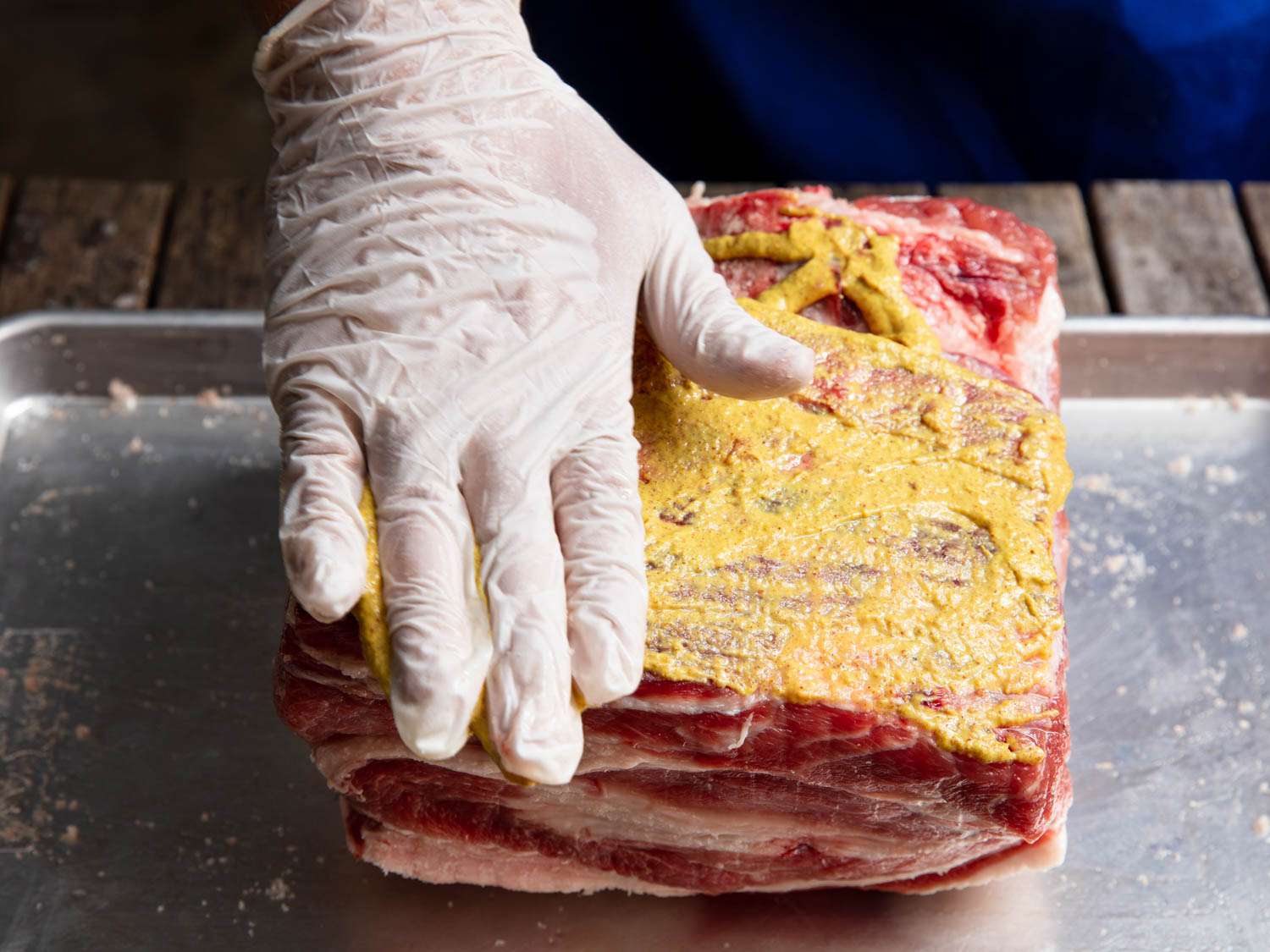 The pork shoulder is daintily smeared with mustard by a gloved hand.
