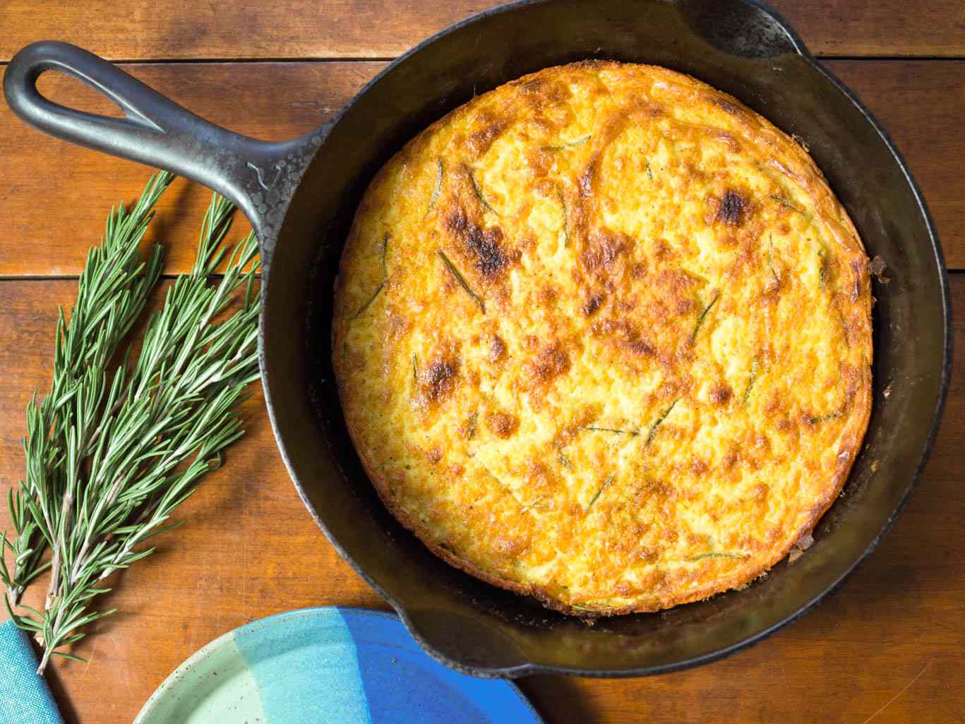 A cooked farinata in a cast iron skillet next to two sprigs of rosemary on a wooden surface. 