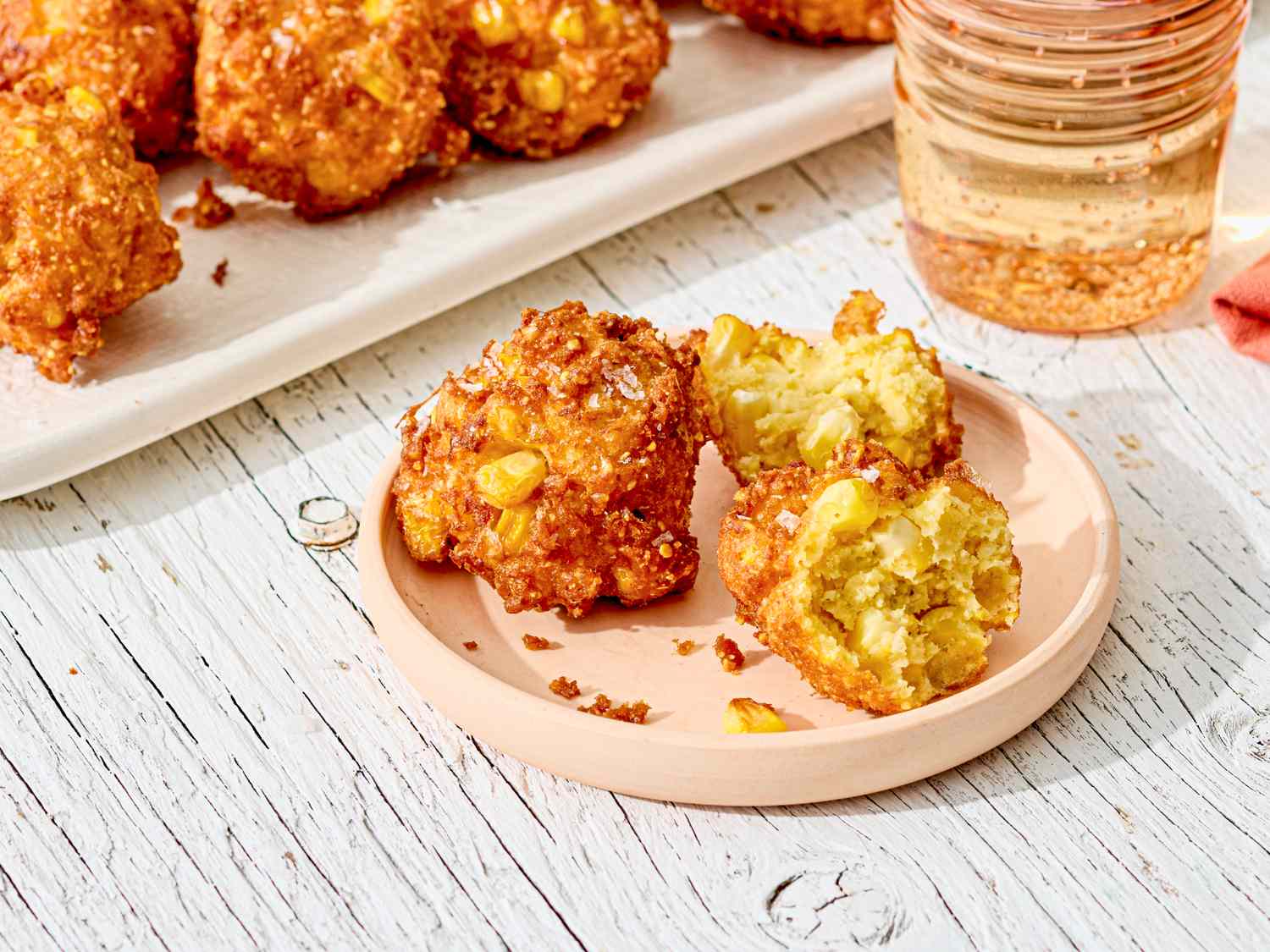 Plate of southern corn fritters nearby a tray of fritters and a glass of beverage