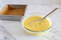 A whisk in a Pyrex bowl of batter next to a baking pan lined with parchment paper