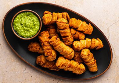 An oval black ceramic plate holding a pile of golden brown tequeÃ±os and a small bowl holding the avocado dipping sauce called guasacaca.