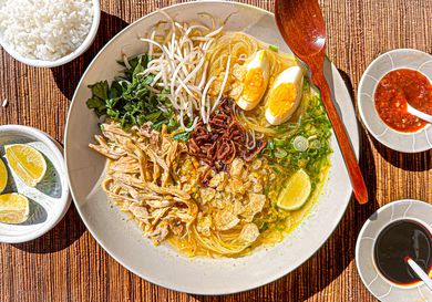 Bowl of Soto Ayam with wooden spoon, surrounded by limes, sambal, soy sauce and rice on a bamboo matt