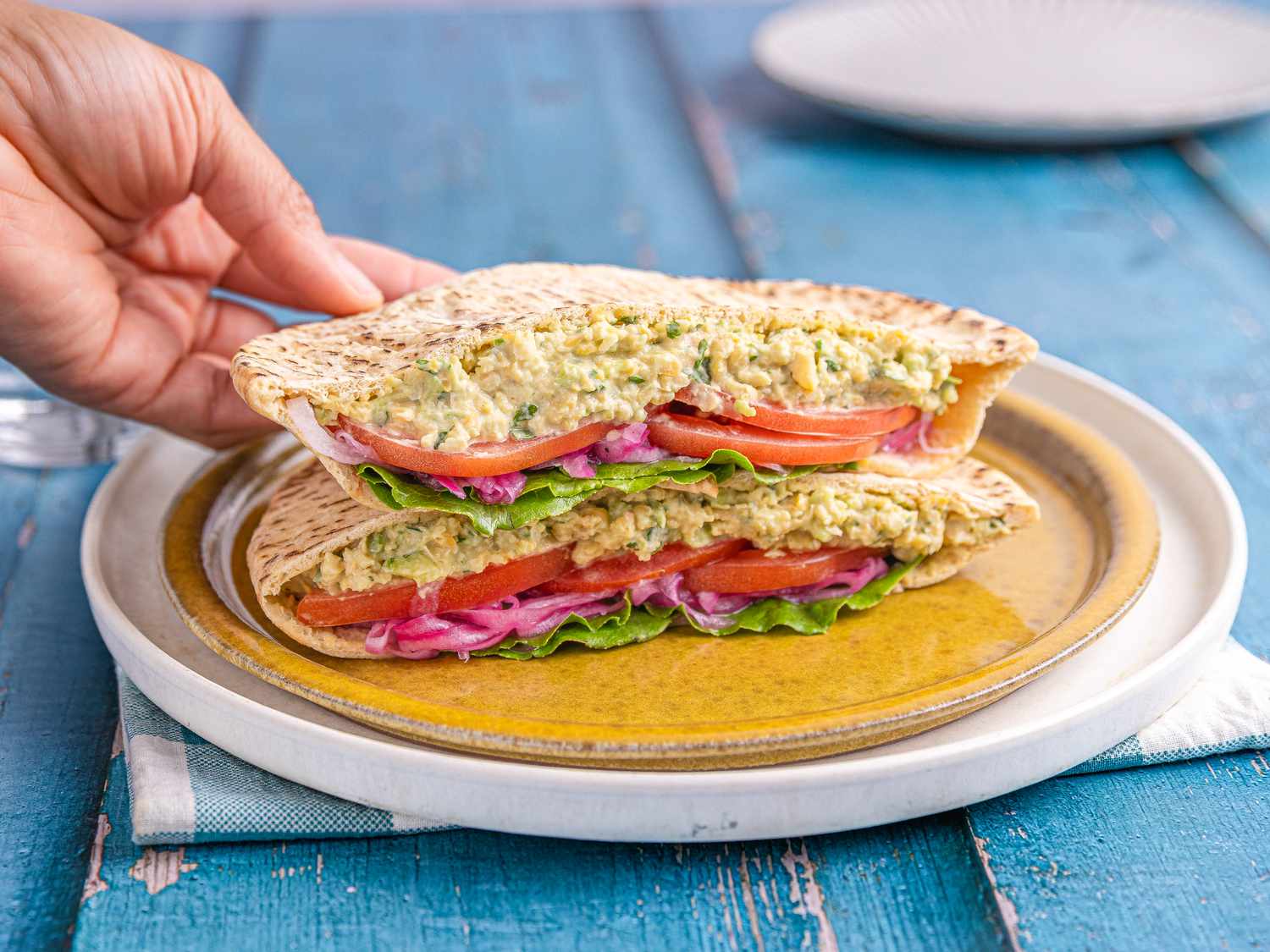 A hand holding a chickpea salad sandwich cut in half, filled with lettuce, red onion, and tomato, placed on a plate
