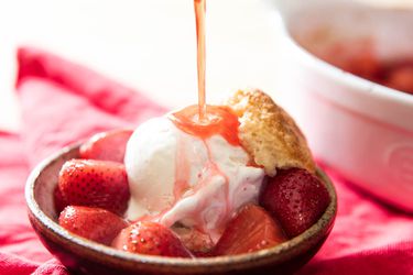 A bowl of roasted strawberries, topped with a scoop of vanilla ice cream and half a biscuit. The bowl is being drizzled with the roasted strawberry pan juices.