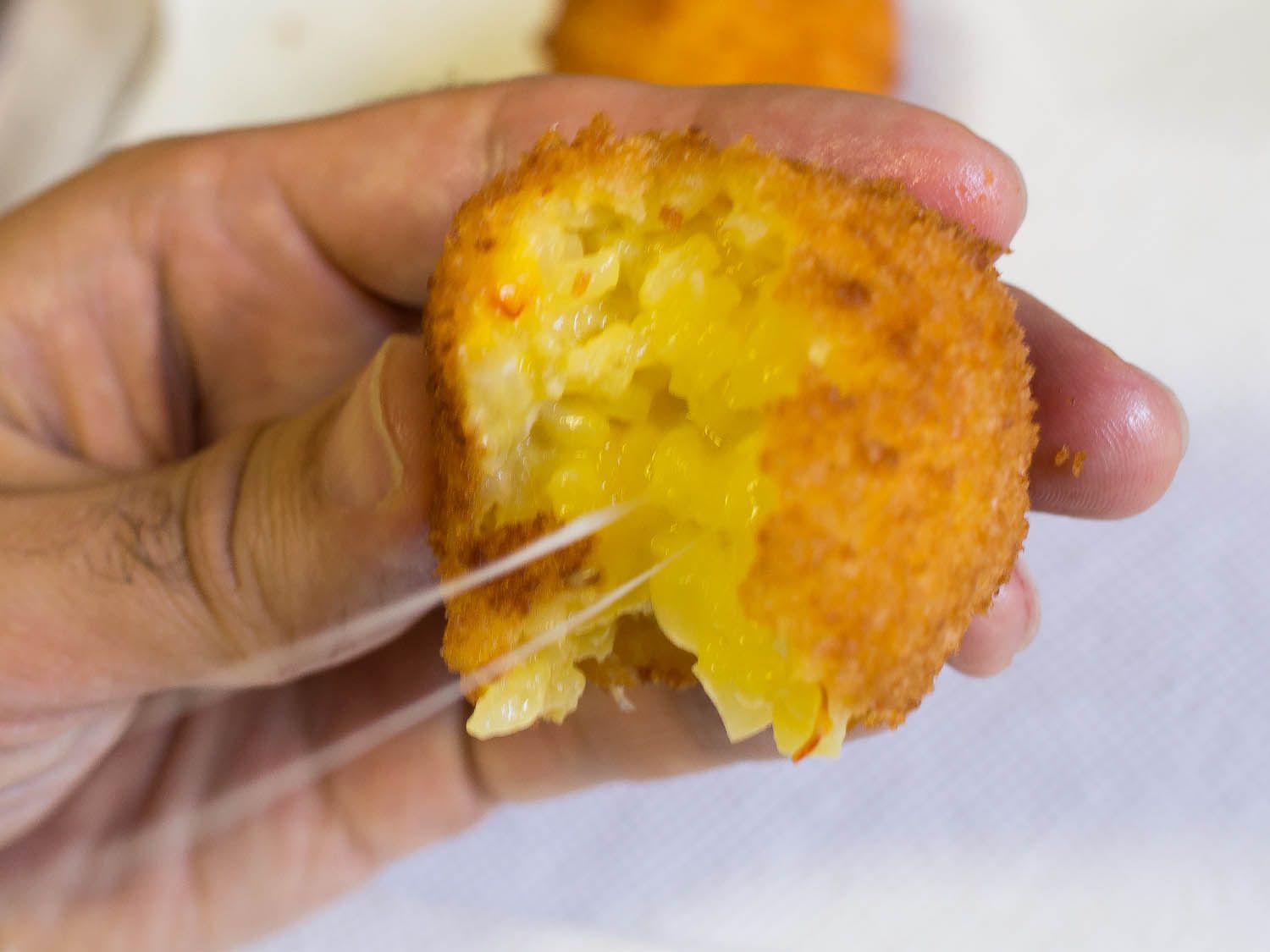 A bitten arancino being pulled away from the off-camera mouth of author. The arancino is trailing several strands of mozzarella.