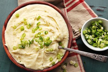 A bowl of creamy mashed potatoes in a red-brown bowl with sliced scallions on top