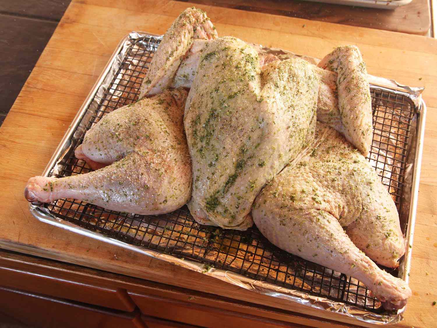 An uncooked, whole, spatchcocked turkey covered in melted herb butter sitting on a wire rack over a foil-covered baking sheet.