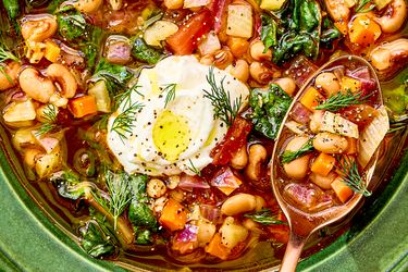 A bowl of stew with blackeyed peas vegetables greens and a dollop of cream topped with dill alongside a spoon