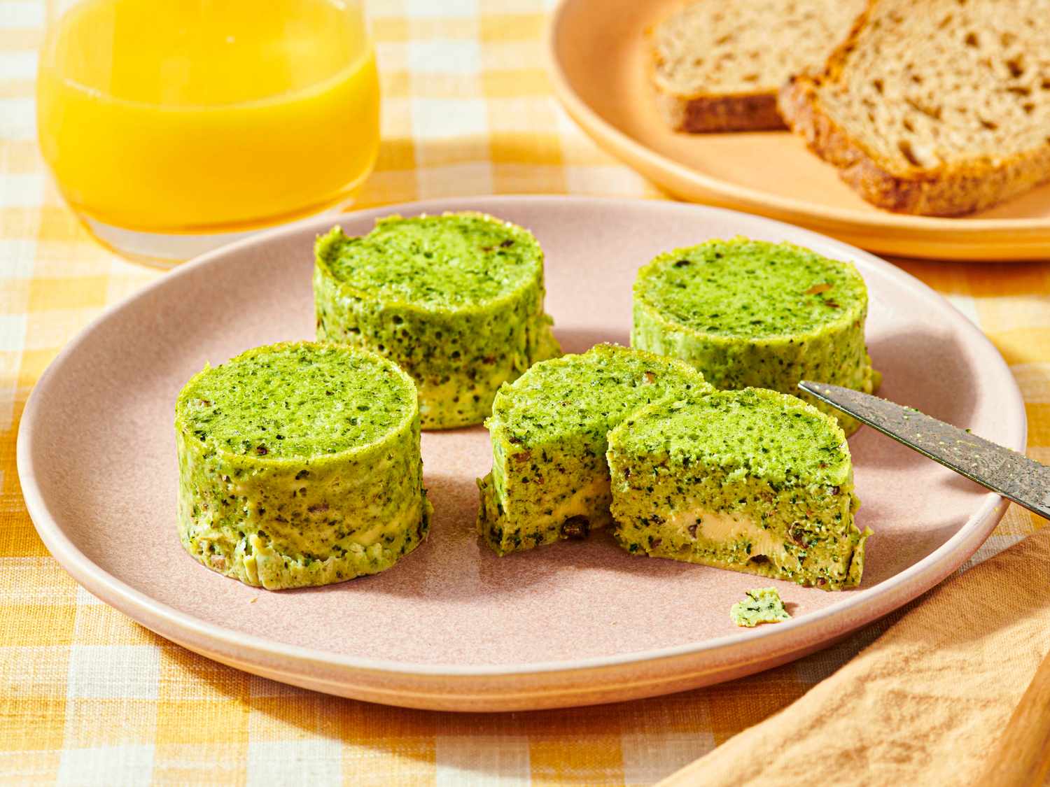 Four round spinach and egg bites on a plate with one cut open accompanied by orange juice and bread in the background
