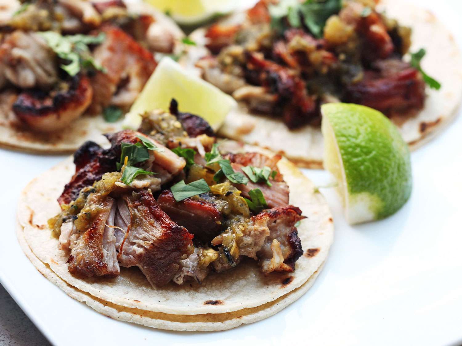 Three sous vide carnitas tacos topped with salsa verde and cilantro, with lime wedges on the side.