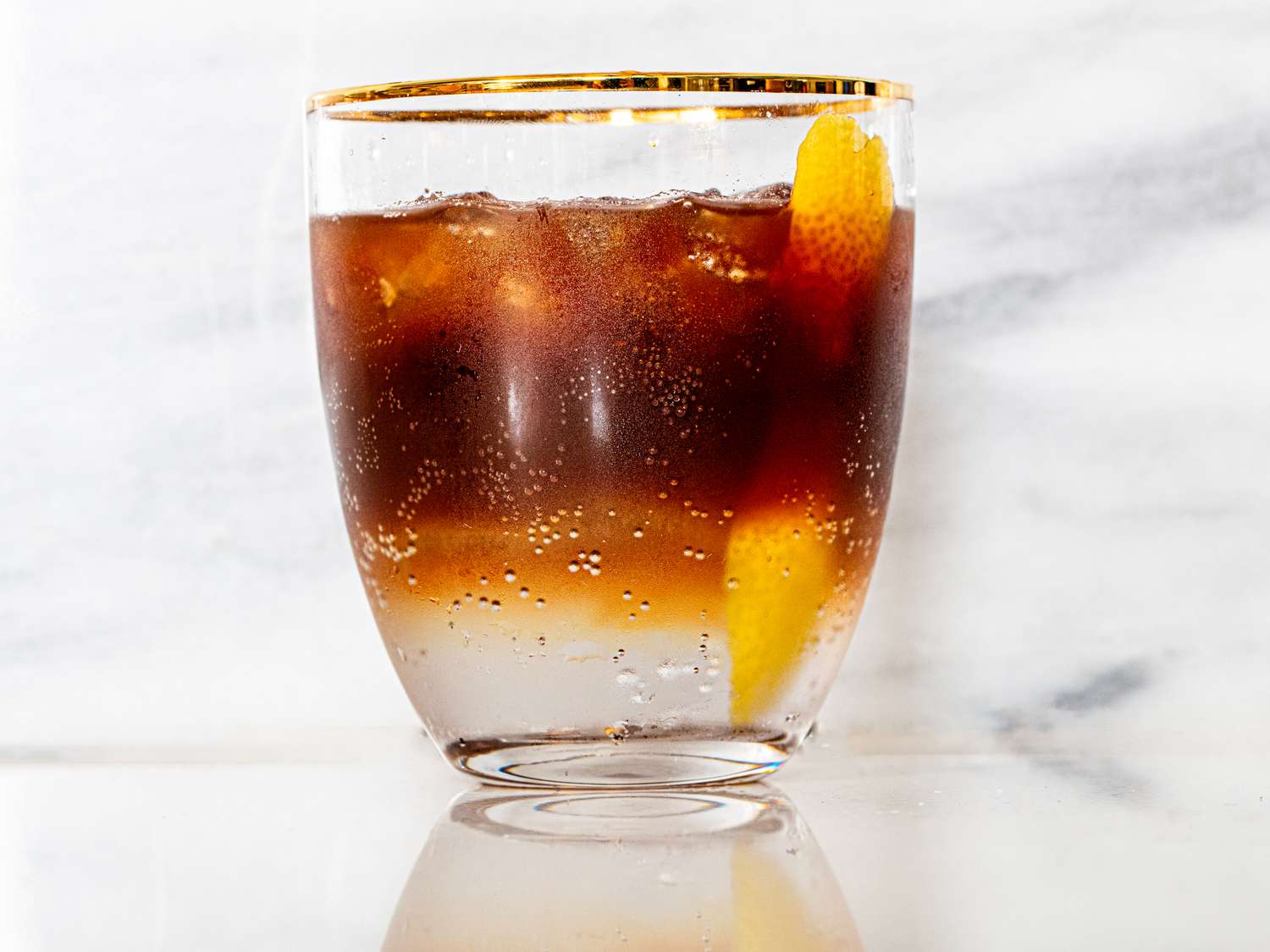 A clear glass filled with a layered drink of espresso on the top and tonic water on the bottom, with a stip of lemon peel in the glass and some ice.