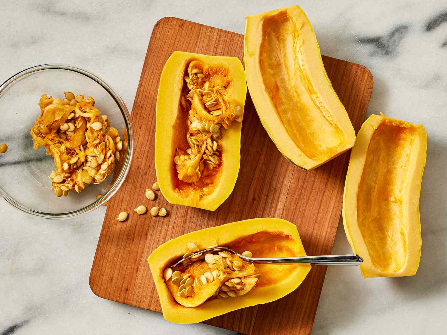 Squash cut into quarters on a wooden board. 2 quartes have the seeds scooped out and put into glass bowl. One quarter has a spoon scooping out seeds, and one still has seeds 
