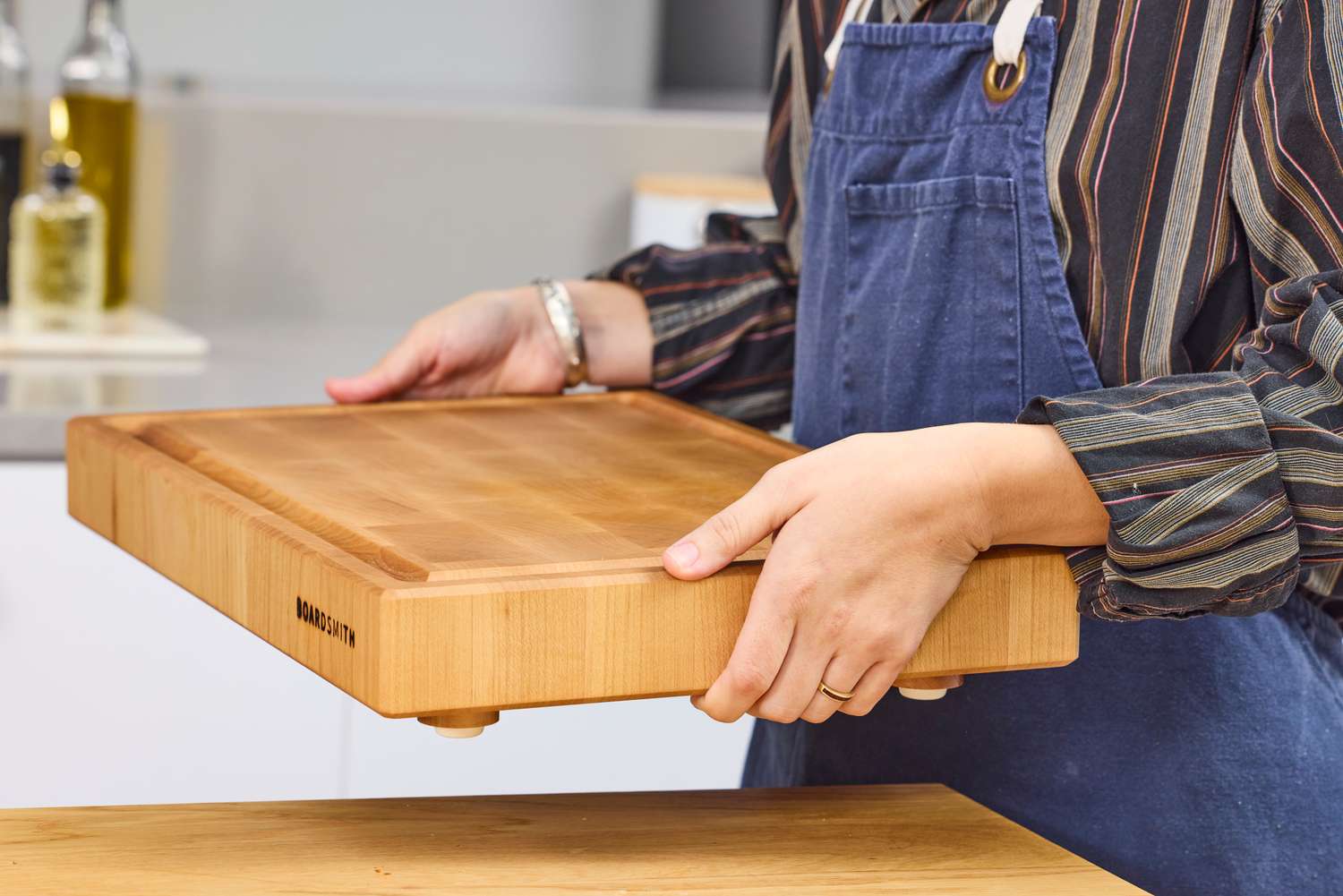 A person holding a Boardsmith Maple End-Grain Cutting Board in a kitchen setting wearing a striped shirt and an apron