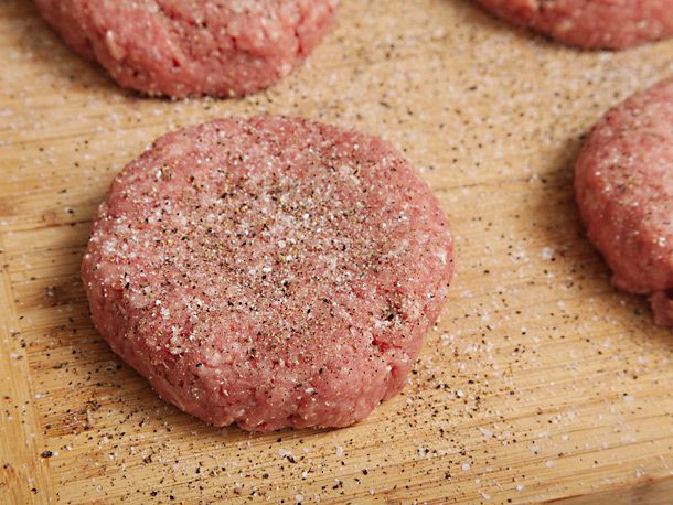Formed burger patties are sprinkled liberally with salt and pepper on a cutting board.