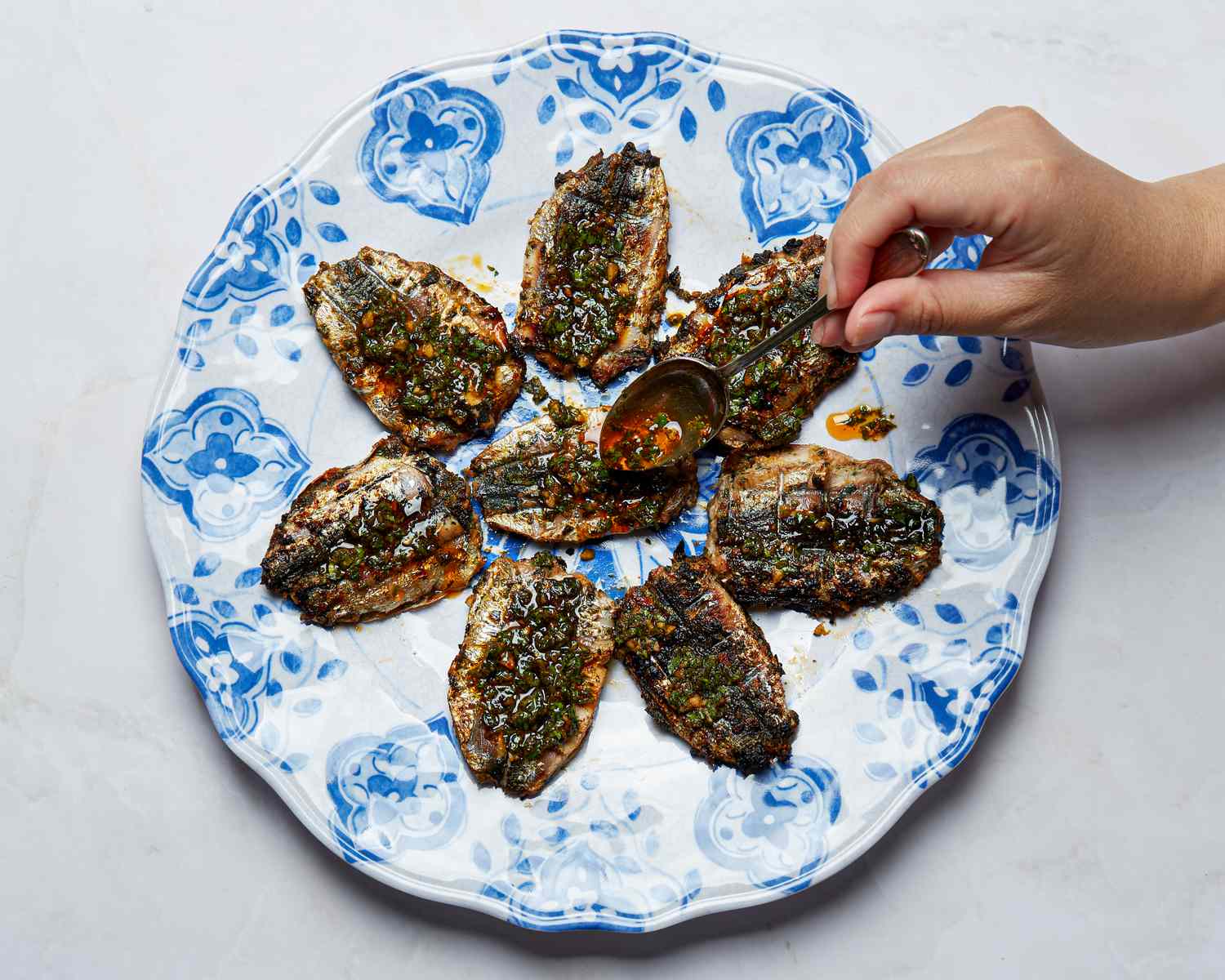 Cooked sardines arranged on a decorative plate with a hand adding sauce to one with a spoon
