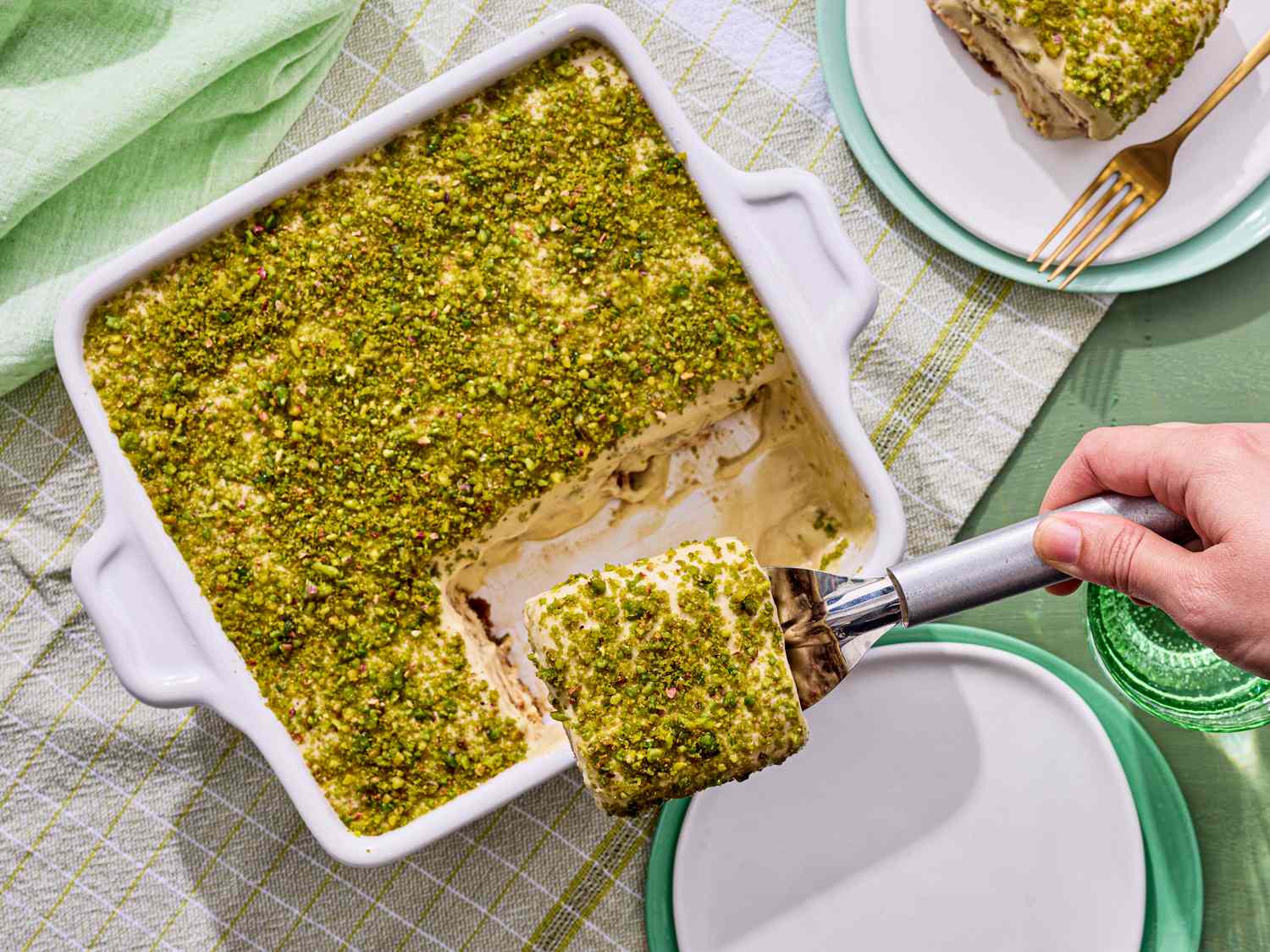 A dish of pistachio tiramisu with a portion being served using a spatula placed on a checkered tablecloth with plates nearby