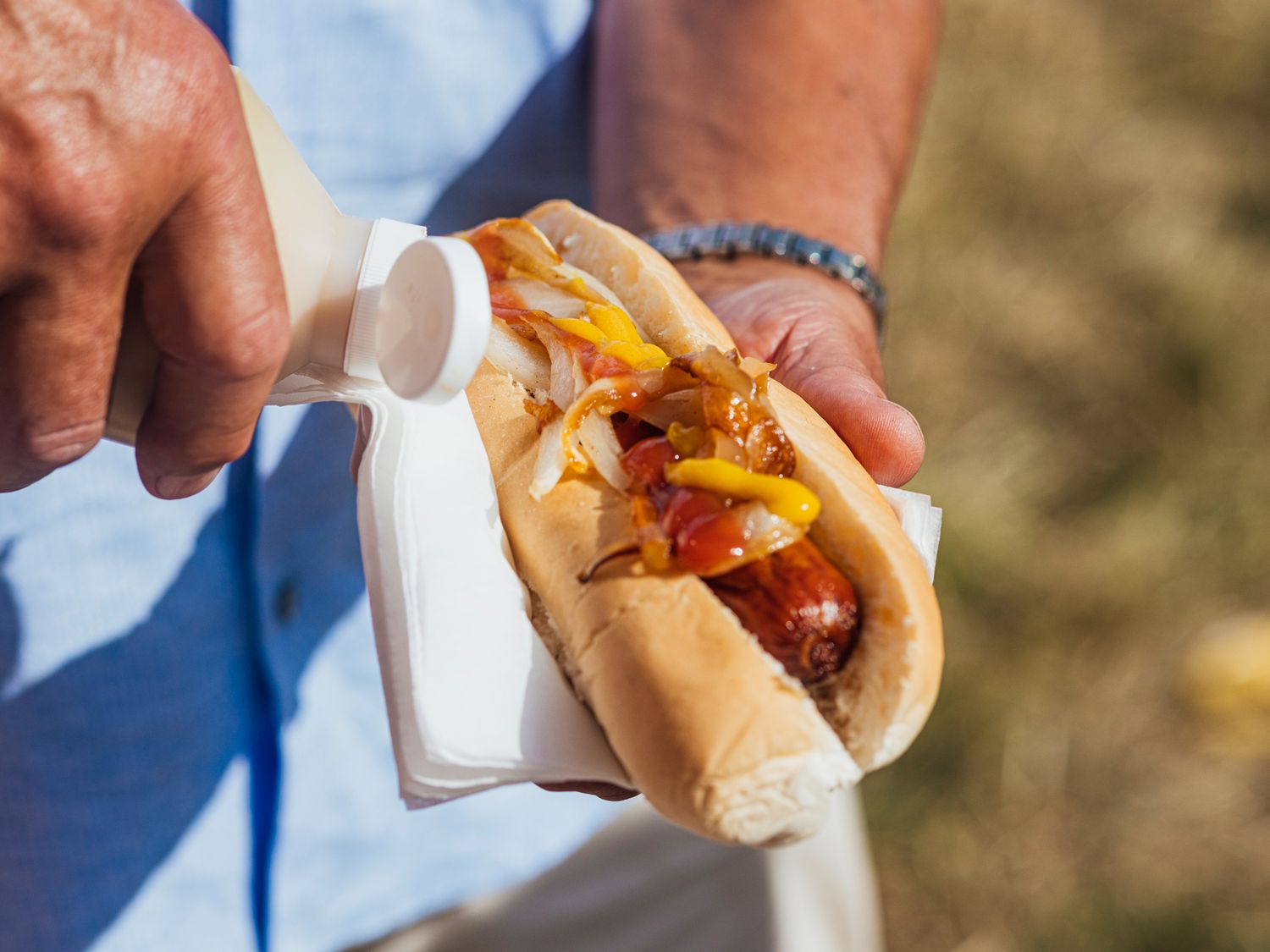 A person applying sauce to a hot dog with onions and mustard on a bun