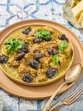 Lamb and prune Tagine with side of couscous, prunes, bread and cutlery on a blue tile surface 
