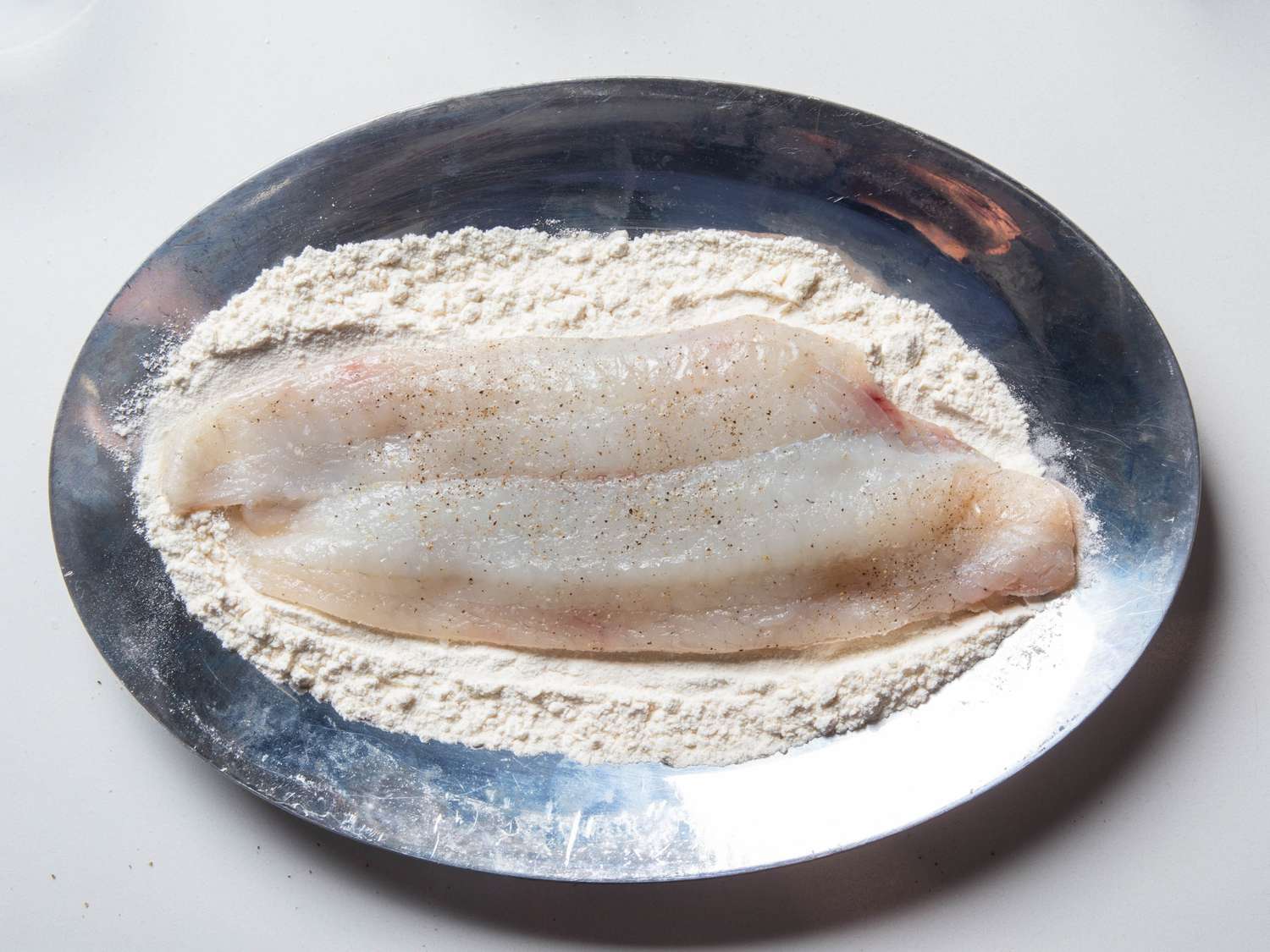 A fish fillet with freshly ground black pepper visible on it sits in a hallow dish of flour, ready for dredging.