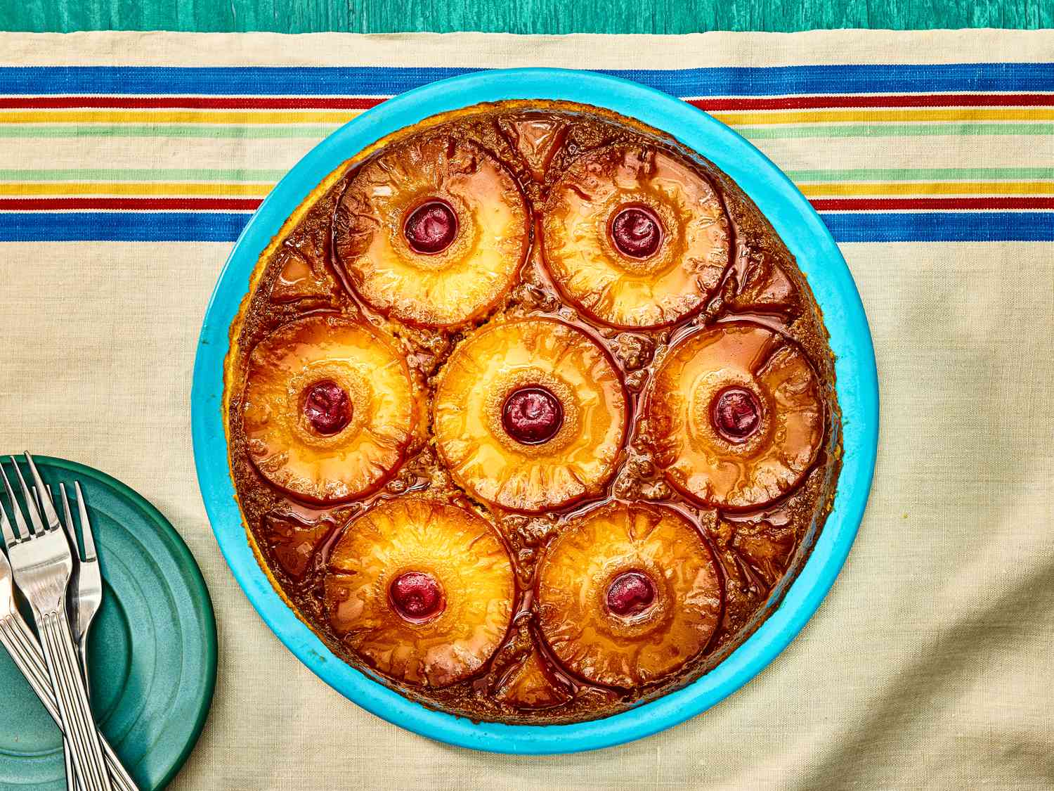 Overhead angle of pineapple upside down cake on a blue plate. The table cloth is a natural tone with blue, red, yellow and green stripes on the top. 