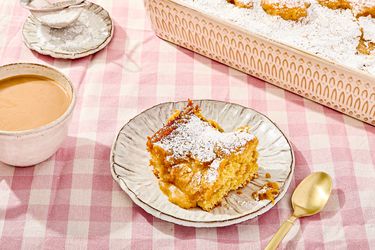 A piece of gooey butter cake on a plate with powdered sugar, with a cup of coffee and a serving tray of cake in the background