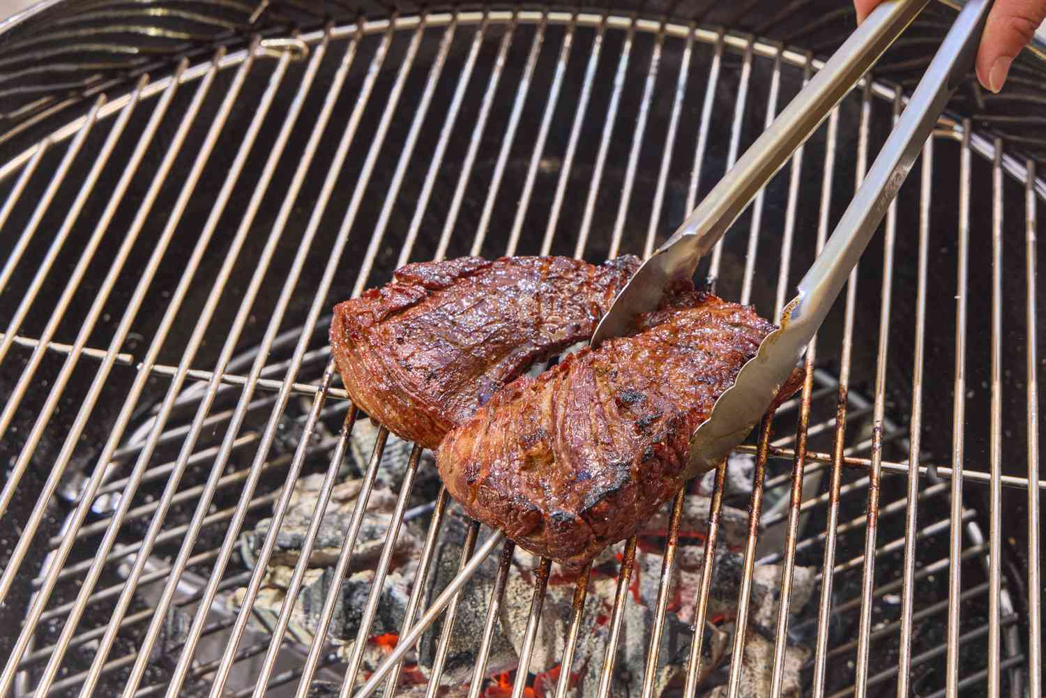 A hand using tongs to move steak on the Weber Original Kettle Premium 22-Inch Charcoal Grill