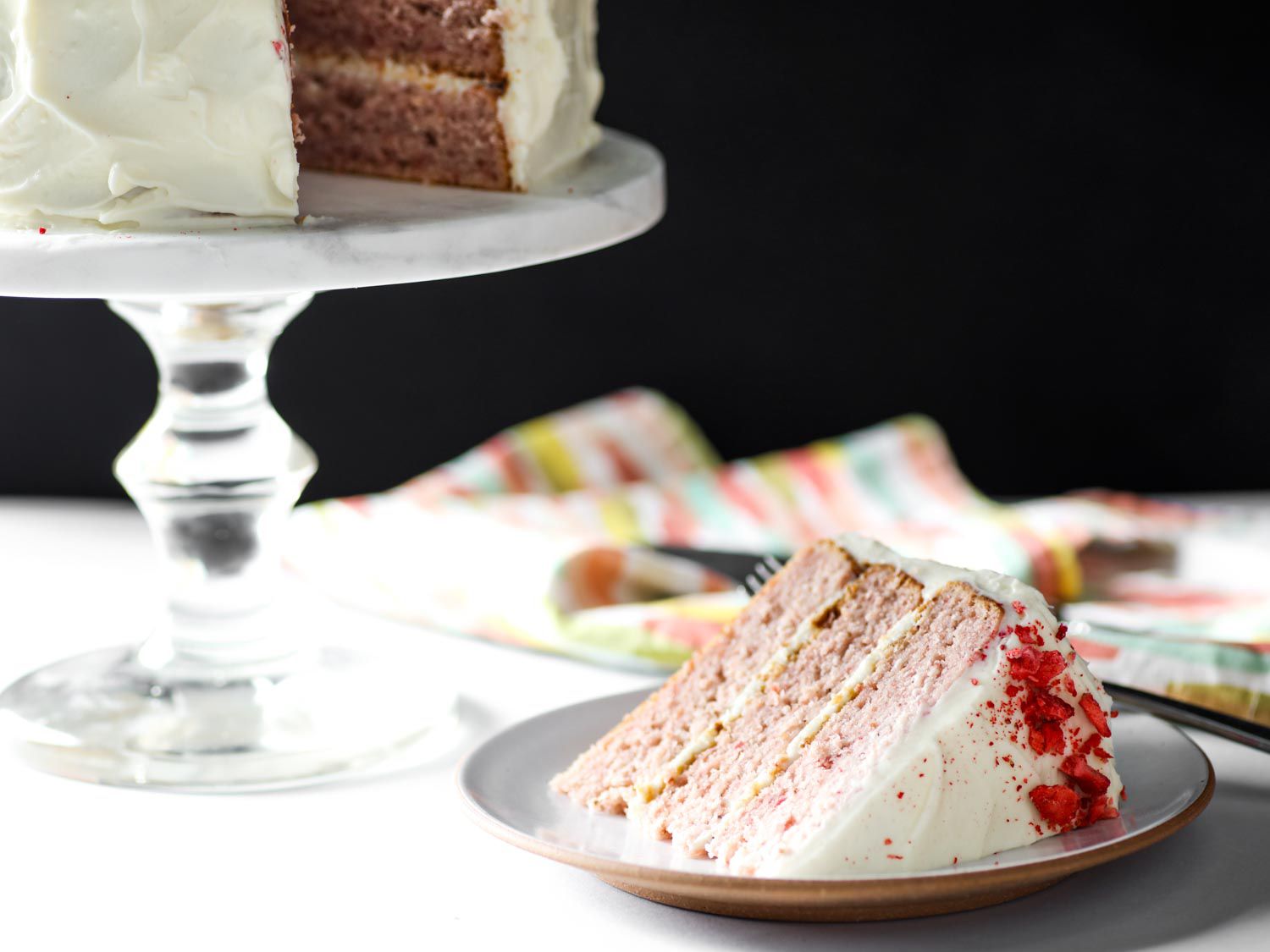 Plated wedge of strawberry layer cake with the rest of the cake on a pedestal in the background.