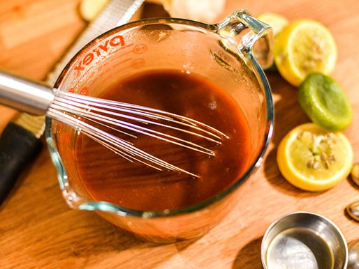 Overhead view of a liquid measuring cup filled with barbecue sauce. A whisk rests against the side of the cup, and cashed lime and lemon halves are strewn to one side.