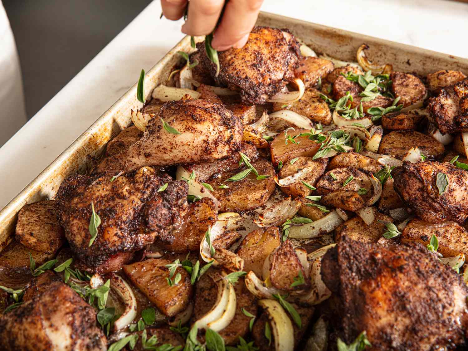 Sheet pan dish with seasoned chicken legs roasted potatoes onions and fresh herbs hand placing chicken on the pan