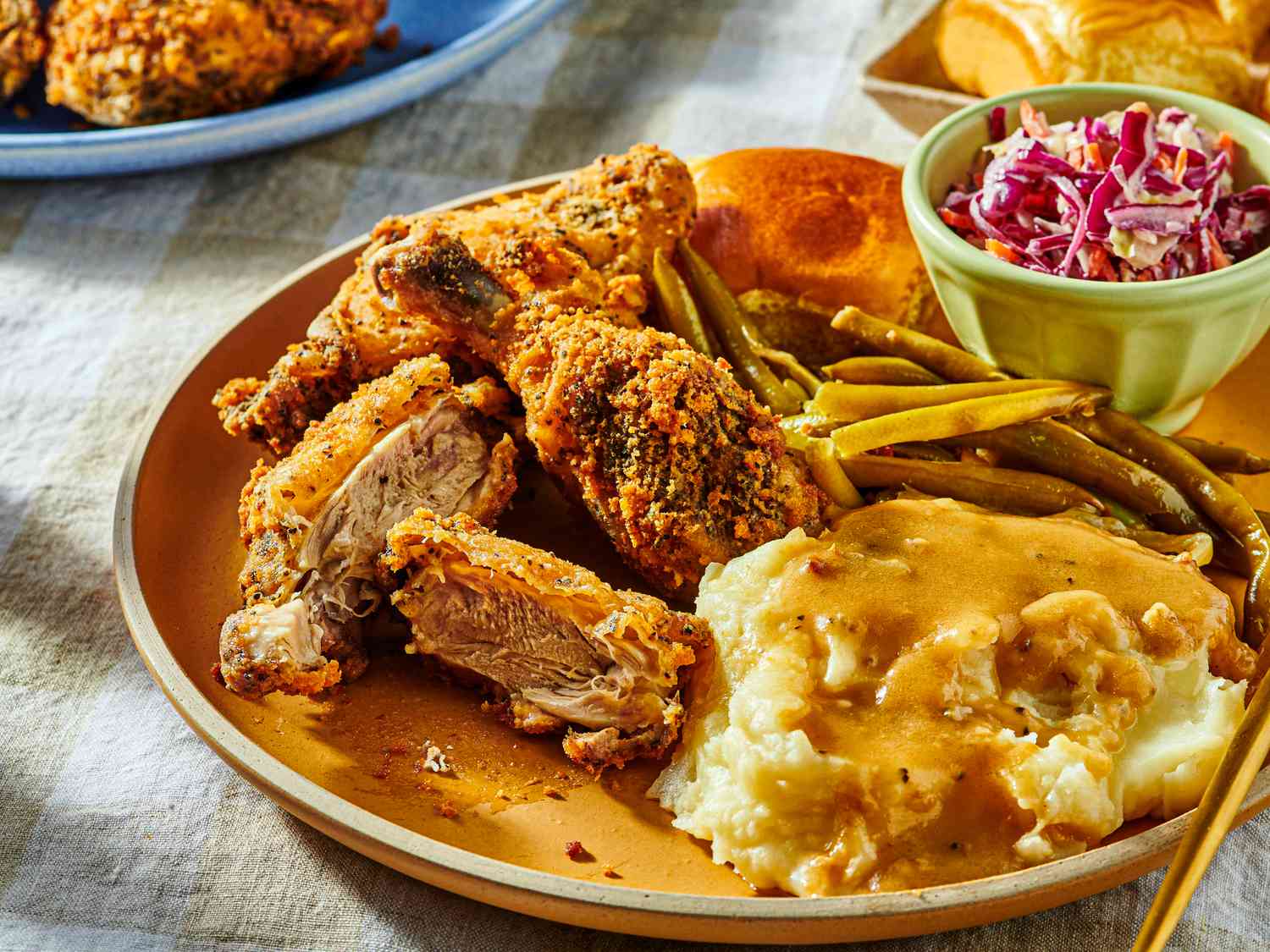 Dish of half eaten fried chicken with coleslaw, greenbeans, and mashed potatoes, with a fork and serving dishes in the background