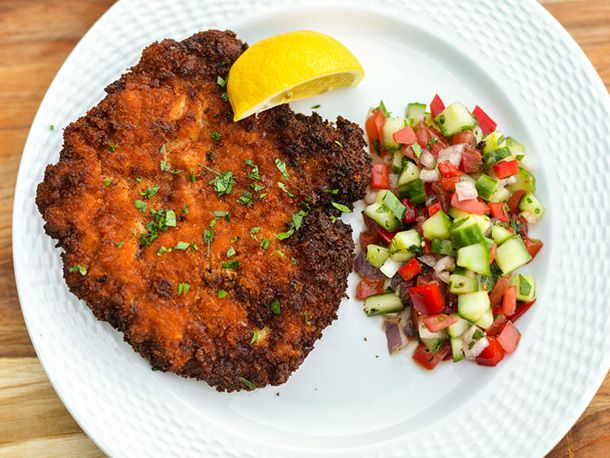 Chicken schnitzel on a white plate with a lemon wedge and a serving of tomato cucumber salad. 