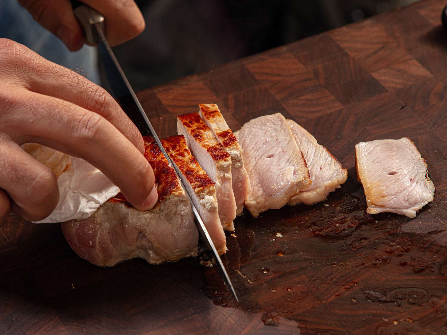 A person slices cooked meat on a wooden cutting board using a knife