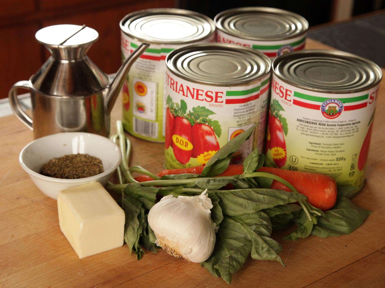 Cans of whole peeled plum tomatoes beside fresh basil, carrot, garlic, butter, dried herbs, and oil dispenser