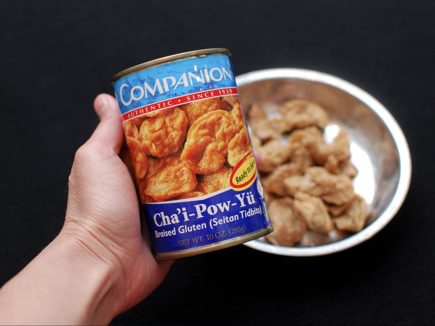 A hand holding a can of seitan tidbits, and some on a plate in the background.