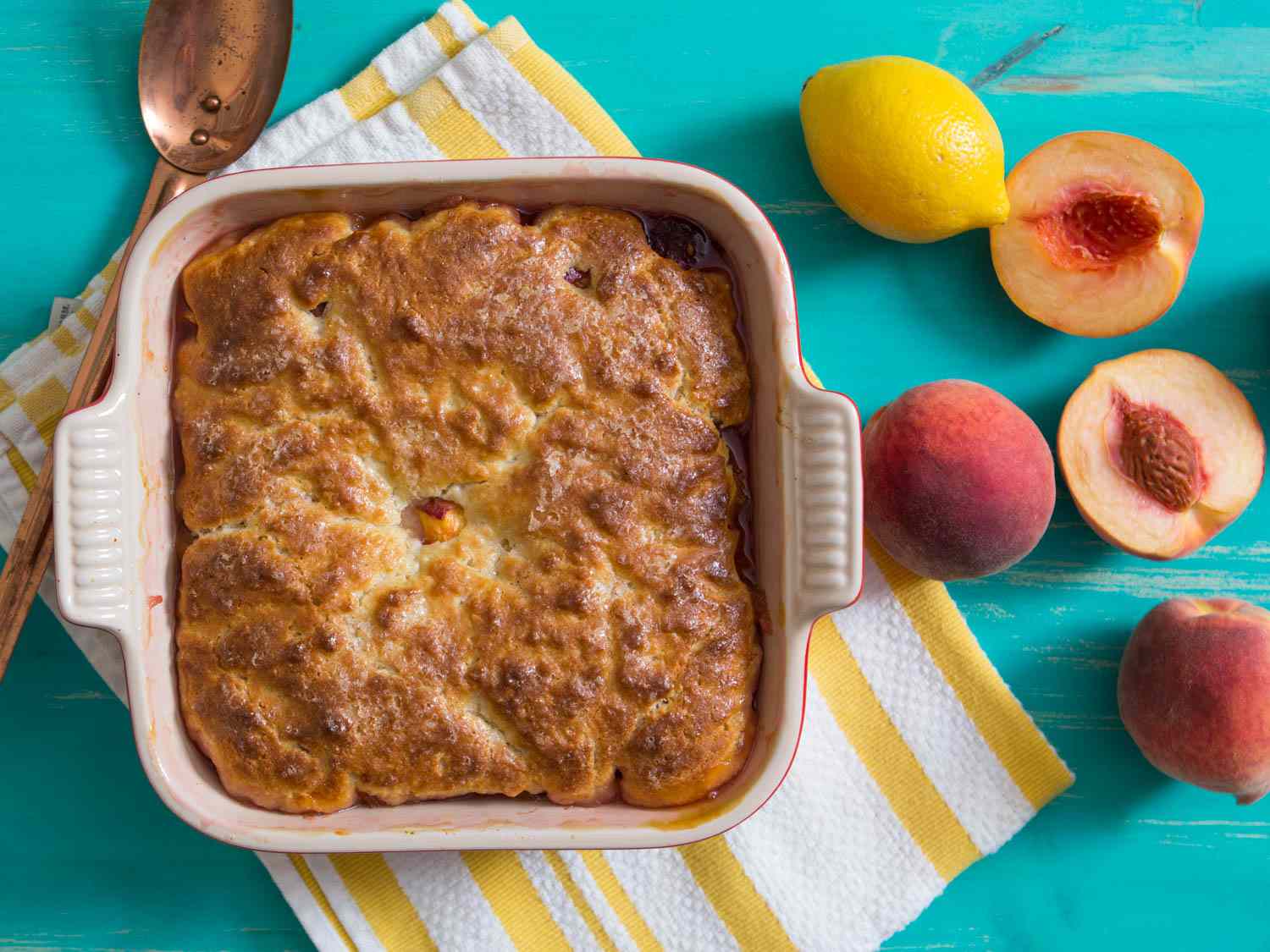 The finished peach cobbler with a golden brown crust, next to a lemon and some split and whole ripe peaches.