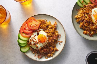 A plate of rice topped with a fried egg on a plate, garnished with cucumber and tomato slices, and showered with fried shallots. There is another plate to the right periphery of the image, as well as several glasses of liquid in the shot.
