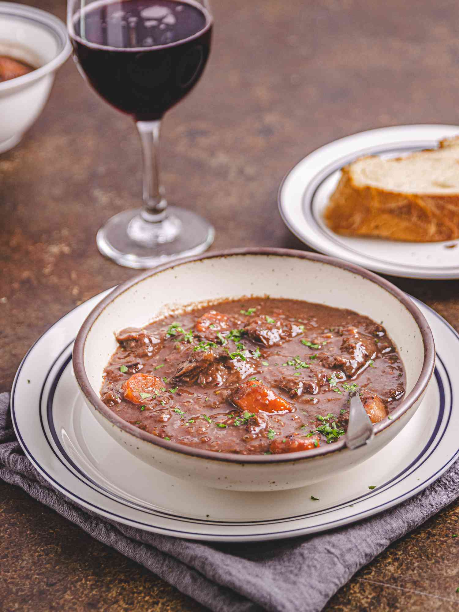 Bowl of stew on a plate on top of a blue napkin, with a glass of red wine, and a piece of bread on the background 