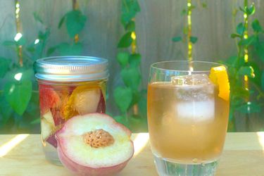 A canning jar of DIY peach liqueur next to a stirred cocktail and a fresh, halved peach.
