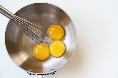 Overhead view of egg yolks sitting in a saucepan. A whisk is balanced on the rim of the pan.