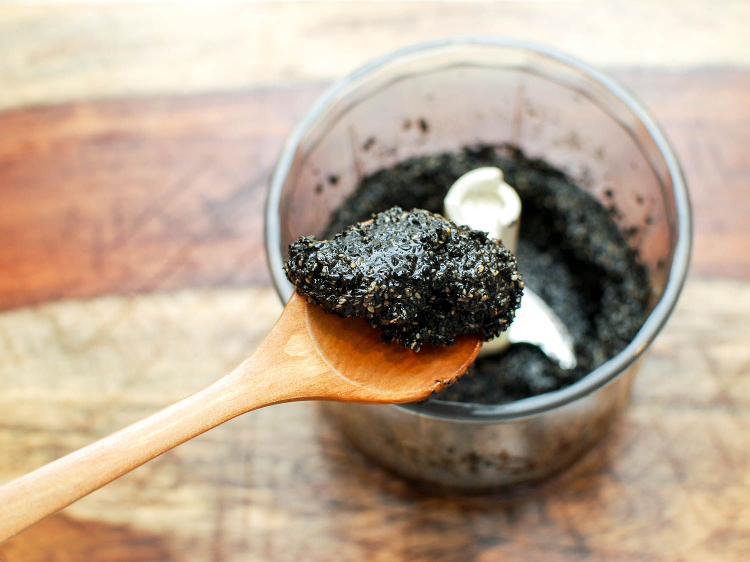 A wooden spoon scoops some ground black sesame paste from a mini food processor.