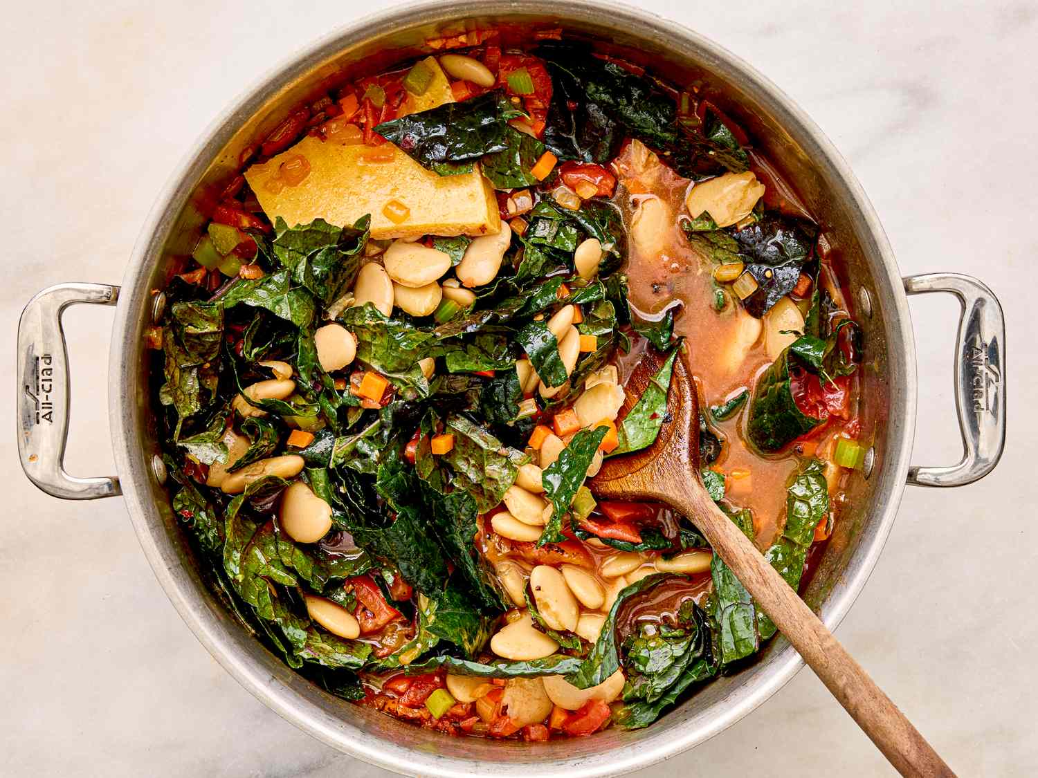 A pot containing a soup made with white beans collard greens and tomato chunks with a wooden spoon inside