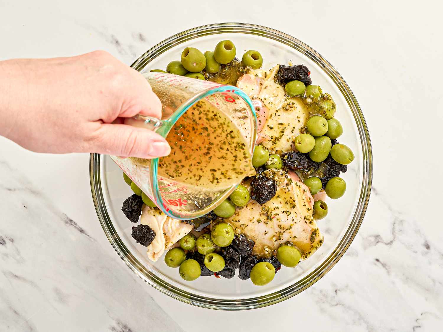 Marinade being poured onto chicken with olives and prunes in a bowl